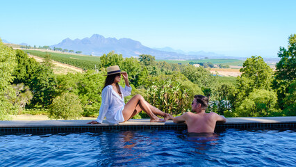 Couple of men and women relaxing at a swimming pool with a view over a Vineyard landscape at sunset with mountains in Stellenbosch, near Cape Town, South Africa.  - obrazy, fototapety, plakaty