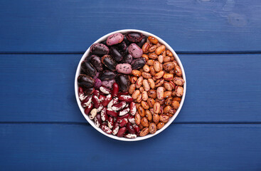 Different kinds of dry kidney beans in bowl on blue wooden table, top view