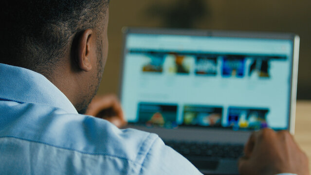Backside unknown man african american businessman web designer employee sit at home office searching films TV series on internet website e-service looking at computer laptop screen over shoulder view