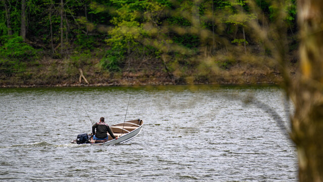 A lone man in a boat seems to be low in the water as he uses his motor to cross the choppy waters of this lake.  Fisherman heading out for a day of fun with fishing poles at the ready.  
