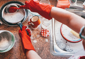 Housewife washing dishes in the kitchen sink. Senior woman washing crockery in front to the window, wearing red protective gloves.