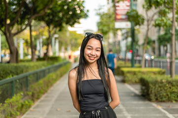 A pretty young lady standing in a park wearing a black strapless tank top and sunglasses, smiling prettily while her hands are behind her back. Trees,bushes and fence are in the background.