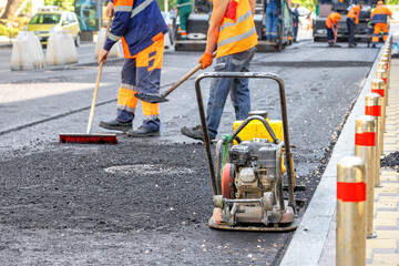 An old worn-out small compacting slab against the backdrop of a team of road workers laying fresh asphalt on a city street in blur. - 604623798