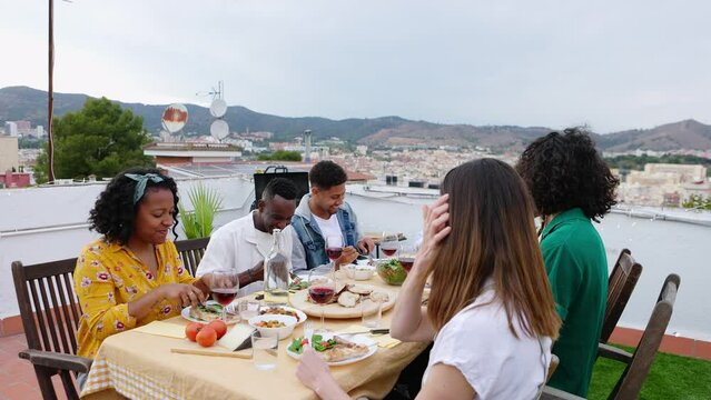 Gathering of young friends having fun at barbecue dinner party on rooftop over Barcelona city background.
