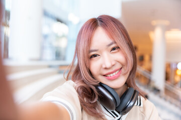 happy female asian student selfie and video conference with her high school friend by technology smart phone and wireless headphone for listen music after test exam in library campus in university