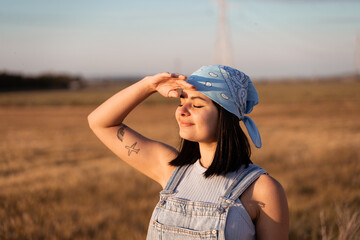 The dark-haired young woman wearing a blue scarf puts her hand on her forehead to block out the sunlight and is in the countryside on a summer afternoon.