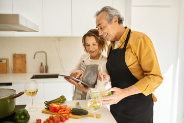 Cheerful senior couple cooking and using digital tablet, looking for nice recipe on Internet in kitchen at home