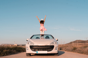 vertical shot European girl dressed in pink outdoors in her convertible car on a highway during the...