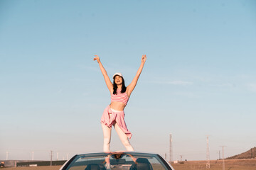 white European girl dressed in summer clothes sits on the seat of her white convertible car and raises her hands to the sky as she rides on a summer afternoon.