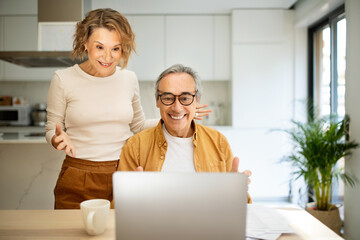 Happy senior spouses man and woman sitting at kitchen table in front of laptop computer, looking at screen and smiling