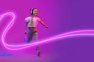Fototapeta na wymiar Cheery little girl using wireless headphones, running in neon light