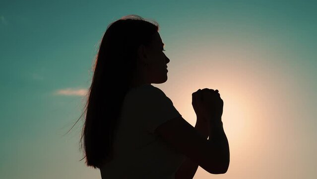 Christian woman praying at sunset close-up. Girl on background of sky in rays of sun prayer to family and children. Relaxation and meditation in nature. Faith in god. Apologize. Hope. Silhouette
