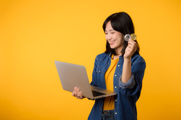 Happy young asian woman wearing yellow t-shirt denim shirt holding digital crypto currency coin and laptop isolated on yellow background. Digital currency financial concept.