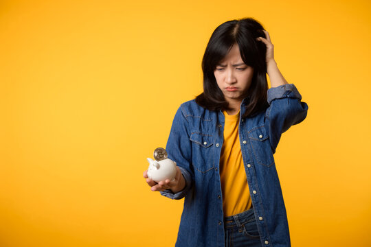 Unhappy asian young woman wearing yellow t-shirt denim shirt pulling digital coin crypto currency out of piggy bank isolated on yellow background. Payment digital money debt financial concept.