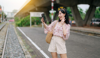 Asian young woman traveler with weaving basket using a mobile phone beside railway train station in Bangkok. Journey trip lifestyle, world travel explorer or Asia summer tourism concept.
