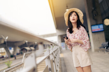 Asian young woman traveler with weaving basket using a mobile phone beside railway train station in Bangkok. Journey trip lifestyle, world travel explorer or Asia summer tourism concept.
