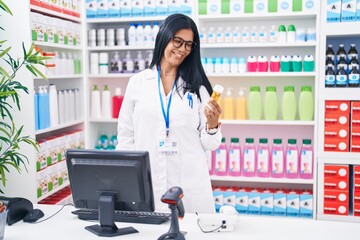 Middle age hispanic woman pharmacist using computer holding pills bottle at pharmacy