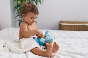 Adorable hispanic baby sitting on bed playing with elephant doll at bedroom