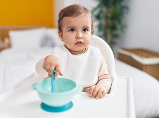 Adorable hispanic baby holding spoon sitting on highchair at bedroom
