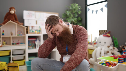 Young redhead man preschool teacher stressed sitting on chair at kindergarten