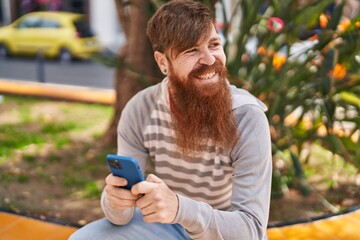Young redhead man using smartphone sitting on bench at park