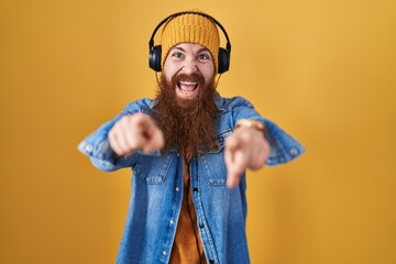 Caucasian man with long beard listening to music using headphones pointing to you and the camera with fingers, smiling positive and cheerful