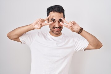 Fototapeta na wymiar Handsome hispanic man standing over white background doing peace symbol with fingers over face, smiling cheerful showing victory