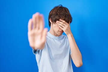 Hispanic young man standing over blue background covering eyes with hands and doing stop gesture with sad and fear expression. embarrassed and negative concept.