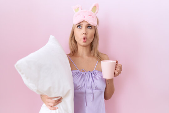Young caucasian woman wearing pajama hugging pillow and drinking coffee making fish face with mouth and squinting eyes, crazy and comical.