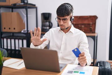 Young hispanic man working using computer laptop holding credit card doing stop sing with palm of...