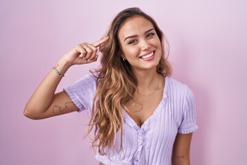 Young hispanic woman standing over pink background smiling pointing to head with one finger, great...