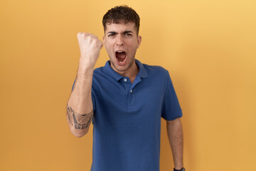 Young hispanic man standing over yellow background angry and mad raising fist frustrated and furious while shouting with anger. rage and aggressive concept.