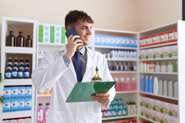 Young hispanic man pharmacist talking on smartphone reading document at pharmacy