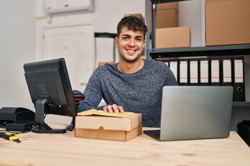 Young hispanic man ecommerce business worker using laptop holding packages at office