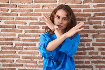 Beautiful brunette woman standing over bricks wall rejection expression crossing arms doing negative sign, angry face