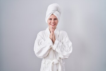 Blonde caucasian woman wearing bathrobe praying with hands together asking for forgiveness smiling confident.