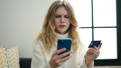 Young blonde woman using smartphone and credit card sitting on sofa at home