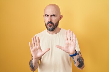Hispanic man with tattoos standing over yellow background moving away hands palms showing refusal and denial with afraid and disgusting expression. stop and forbidden.