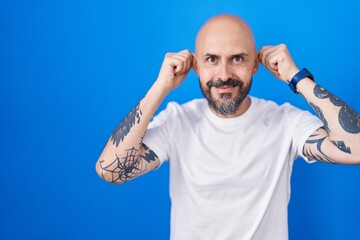 Hispanic man with tattoos standing over blue background smiling pulling ears with fingers, funny...