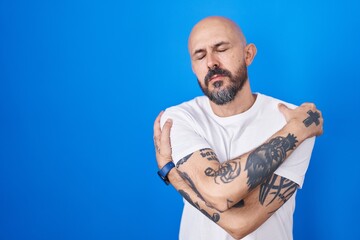 Hispanic man with tattoos standing over blue background hugging oneself happy and positive, smiling confident. self love and self care