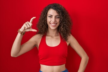 Hispanic woman with curly hair standing over red background smiling and confident gesturing with hand doing small size sign with fingers looking and the camera. measure concept.