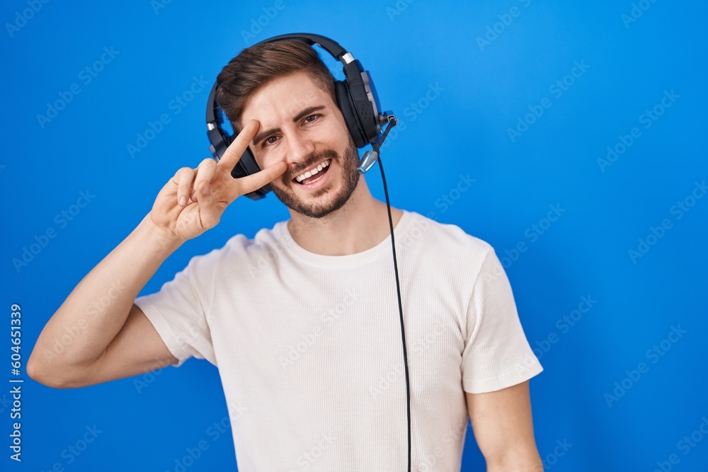 Poster Hispanic man with beard listening to music wearing headphones doing peace symbol with fingers over face, smiling cheerful showing victory