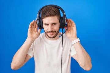 Hispanic man with beard listening to music wearing headphones trying to hear both hands on ear gesture, curious for gossip. hearing problem, deaf