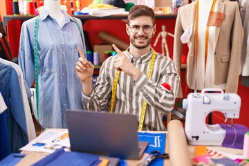 Hispanic man with beard using laptop at tailor room smiling and looking at the camera pointing with two hands and fingers to the side.