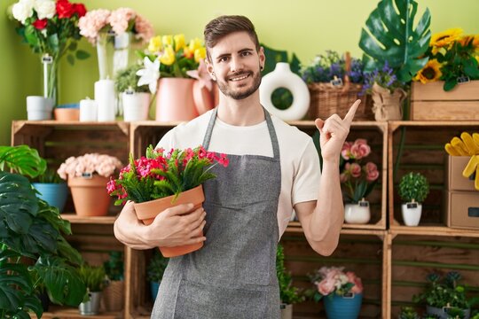 Hispanic man with beard working at florist shop holding plant smiling happy pointing with hand and finger to the side