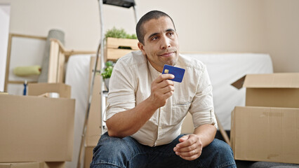 Obraz na płótnie Canvas Young hispanic man holding credit card sitting on sofa thinking at new home
