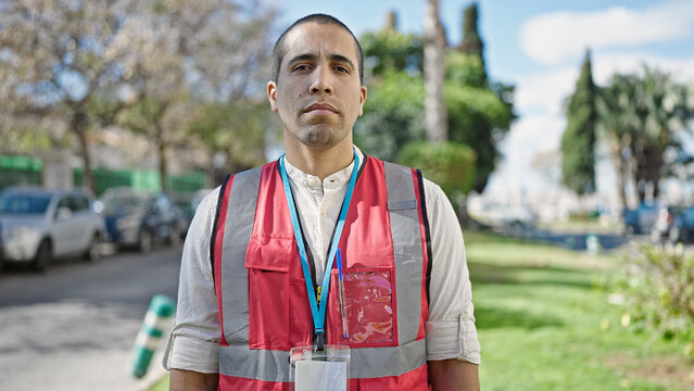 Young hispanic man volunteer wearing vest looking serious at park