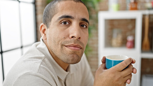 Young hispanic man drinking coffee sitting on table at dinning room