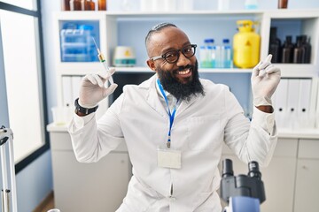 African american man working at scientist laboratory holding syringe smiling happy pointing with hand and finger to the side