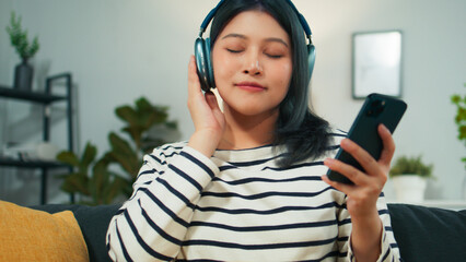 Attractive Asian woman using smartphone listen to podcast music with hand holding wireless headphone. Young female resting stress relief with chilling happy peaceful face sits on sofa in living room